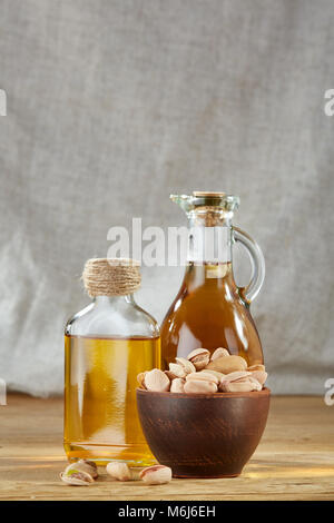 Olio aromatico in un vasetto di vetro e bottiglia trasparente con pistacchio in ceramica ciotola sul tavolo di legno su sfondo chiaro, primo piano. Ancora vita compositi Foto Stock