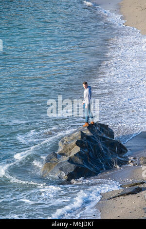 Un giovane uomo in piedi sulle rocce come la marea avanza sul piccolo Fistral Beach in Newquay Cornwall. Foto Stock