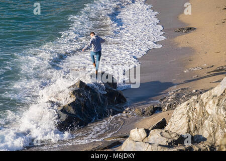 Un giovane uomo di equilibratura su rocce come la marea arriva a poco Fistral Beach in Newquay Cornwall. Foto Stock