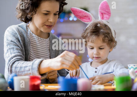 Ritratto di carino piccolo ragazzo indossa orecchie di coniglietto di pittura delle uova di Pasqua in casa con la mamma mentre si prepara per la vacanza con la mamma Foto Stock