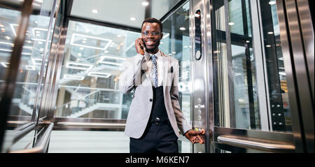 Imprenditore afroamerican in moderno ascensore di vetro parlando al telefono Foto Stock