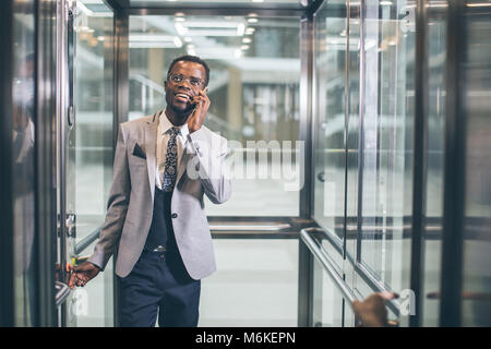 Imprenditore afroamerican in moderno ascensore di vetro parlando al telefono Foto Stock