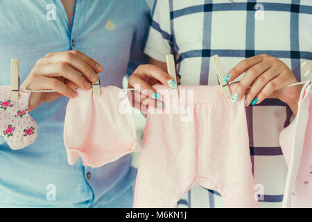 Close-up shot dei futuri genitori appendere abiti rosa per il loro bambino neonato sulla stendibiancheria utilizzando i vestiti-PEG. Nessun volto. Vista delle mani. Foto Stock