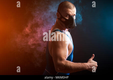 Un forte dai capelli scuri in sportman un lottatore di wrestling blu collant e maschera per la formazione permanente e la mostra il pollice in su contro un blu e rosso vape backgroun di fumo Foto Stock