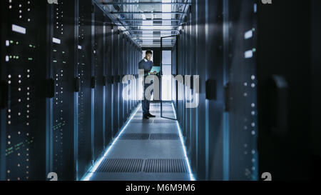 Colpo lungo del tecnico che lavora sul computer portatile in grandi data center pieno di server Rack. Egli esegue il programma di diagnostica e manutenzione, Sistema di set up. Foto Stock