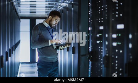 Tecnico IT funziona su computer portatile accanto a un server cabinet in grandi data center. Egli esegue il programma di diagnostica e manutenzione, Sistema di set up. Foto Stock