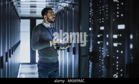 Tecnico IT funziona su computer portatile accanto a un server cabinet in grandi data center. Egli esegue il programma di diagnostica e manutenzione, Sistema di set up. Foto Stock