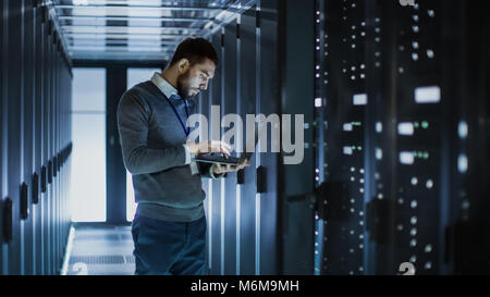 Tecnico IT funziona su computer portatile accanto a un server cabinet in grandi data center. Egli esegue il programma di diagnostica e manutenzione, Sistema di set up. Foto Stock