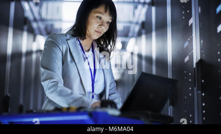 Asian femmina tecnico che lavora su un computer portatile sul Tool Cart ella esegue la scansione dei dischi rigidi. Lei è in un grande centro dati pieno di server Rack. Foto Stock