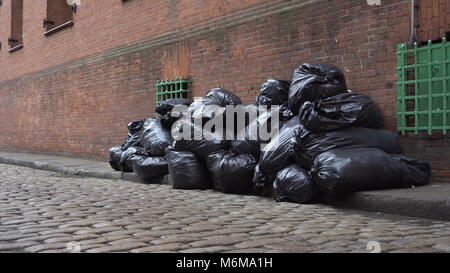 Cestino nero sacchi riempiti con garbage sedersi sul cordolo di Brooklyn, New York Street lungo il lato di un edificio in mattoni in attesa di essere raccolti e smaltiti b Foto Stock