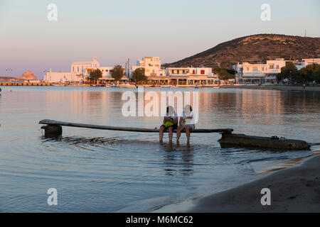 Vista lungo la spiaggia con 2 donne sedeva sul molo al tramonto, Pollonia Milos, Cicladi, il Mare Egeo e le isole greche; Grecia; l'Europa Foto Stock