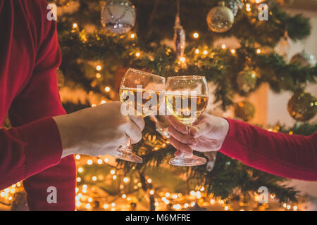 Close up di maschio e femmina mani rasserenanti e azienda bicchieri di vino nella parte anteriore del albero di Natale decorato. Capodanno celebrazione. Foto Stock