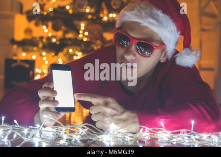 Considerato l uomo con Santa hat sdraiato sul pavimento e lo smartphone che mostra una schermata vuota. Albero di natale in background Foto Stock