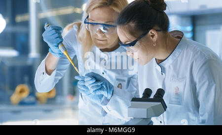 Medical Research Scientist gocce di campione sulla slitta e il suo collega lo esamina al microscopio. Essi lavorano in un moderno laboratorio. Foto Stock