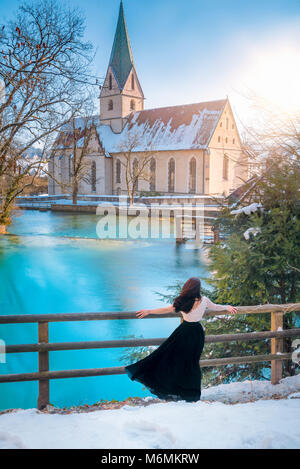 Vista posteriore di un attraente giovane donna vestita elegantemente, ammirando le acque blu della molla di Blautopf, in una giornata di sole, a Blaubeuren, Germania. Foto Stock