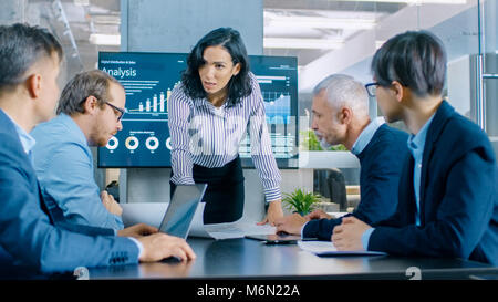 Chief Executive femmina si appoggia e diffonde un progetto Blueprint sul tavolo mostrando loro ai suoi colleghi. Sullo sfondo i grafici a torta e grafici Foto Stock