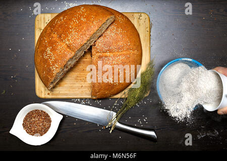 Fatto in casa pane integrale con semi di lino, farina integrale, aneto e coltello su un tavolo di legno. Donna diffondendo la farina in una ciotola. Foto Stock