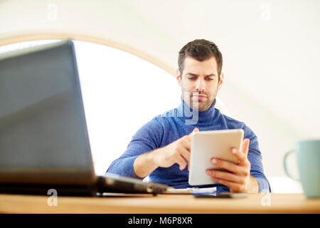 Giovane uomo che indossa un maglione turtleneck mentre usando tavoletta digitale e portatile a casa. Home office. Foto Stock