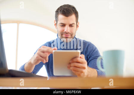 Giovane uomo che indossa un maglione turtleneck mentre usando tavoletta digitale e portatile a casa. Home office. Foto Stock
