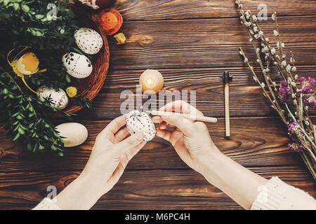 Le mani di pittura delle uova di pasqua con cera d'api vista dall'alto. vacanze di pasqua concetto. Uova, boccioli e fiori, candele e pollo laici piatta con spazio per il testo Foto Stock