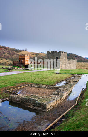 Roman Fort Vindolanda, vista di una ricostruzione di una sezione della torre del Muro di Adriano con scavato serbatoio di acqua in foregound, Northumberland, England, Regno Unito Foto Stock