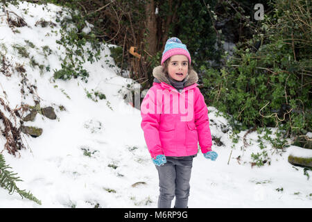 Carino bambina in piedi nella neve indossando giacca rosa e blu inverno pompom hat Foto Stock