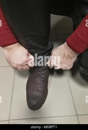 Il tempo di legare shoesThe uomo rannicchiato che si allaccia il brown scarpe chiuse. Sembra che stia indossando pantaloni di pelle e un maglione rosso. Foto Stock