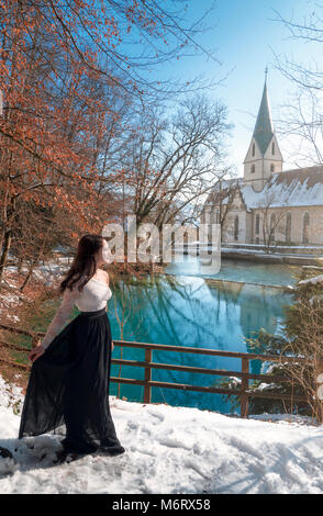 Elegantemente vestito giovane donna bruna, passeggiate attraverso la neve, sulla riva della molla di Blautopf, in una giornata di sole, a Blaubeuren, Germania. Foto Stock