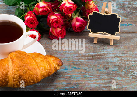 Tazza di tè e caffè con croissant, rose fresche e lavagna con spazio per il testo su un sfondo di legno. Immagine perfetta per la festa della mamma. Foto Stock