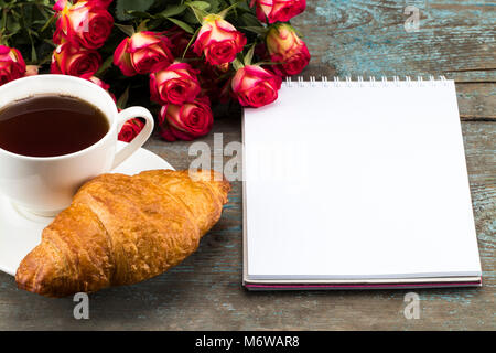Tazza di tè e caffè con croissant, rose fresche , notebook e il foglio di carta su sfondo di legno per la Festa della mamma. Foto Stock