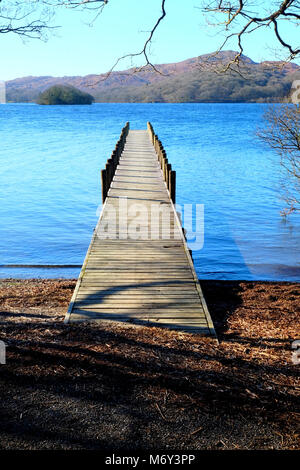 Lungo rettilineo di piede legno pontile proteso oltre un calmo lago blu, con colline verdi campi e boschi della Foresta in background, il sole è shinin Foto Stock