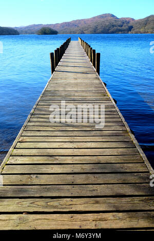 Molto lunghe e diritte simmetrico piede legno pontile proteso oltre un calmo lago blu, con colline verdi campi e boschi della Foresta in background, t Foto Stock