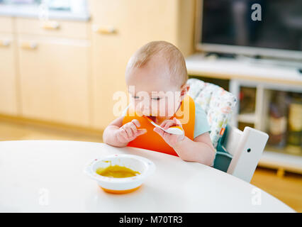 Bambino seduto a pranzare tabella con orange bib e cucchiaio e cercando di mangiare Foto Stock