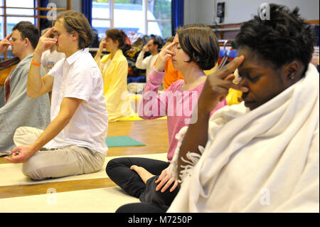 Gruppo di giovani gente mista praticando yogic tecnica di respirazione pranayama chiamato in una hall. Seduti sulle loro Materassini da yoga sul pavimento in parquet. Foto Stock