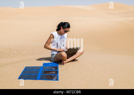 Una donna lavora in remoto sul suo computer tra le dune, alimentato da un pannello solare portatile, autosufficienza e sostenibilità in un ambiente remoto e naturale Foto Stock