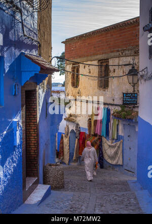 La vista posteriore della donna locale in abito tradizionale djeibella camminando giù colorata strada stretta in scena la città blu di Chefchaouen, Marocco Foto Stock