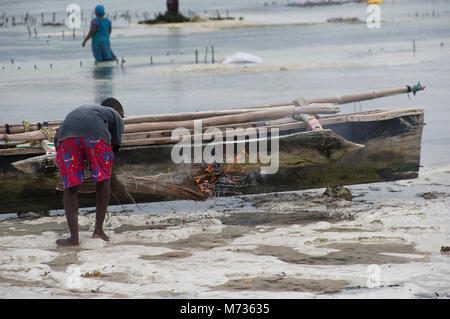 Fisherman utilizzando la masterizzazione di fronde di palma per rimuovere il muschio e verde crescite erbosa dal lato della sua nave da pesca in Jambiani Zanzibar Tanzania Foto Stock