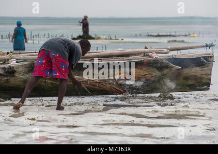 Fisherman utilizzando la masterizzazione di fronde di palma per rimuovere il muschio e verde crescite erbosa dal lato della sua nave da pesca in Jambiani Zanzibar Tanzania Foto Stock