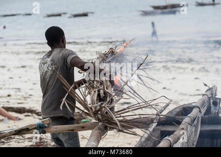 Fisherman utilizzando la masterizzazione di fronde di palma per rimuovere il muschio e verde crescite erbosa dal lato della sua nave da pesca in Jambiani Zanzibar Tanzania Foto Stock