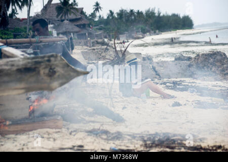 Fisherman utilizzando la masterizzazione di fronde di palma per rimuovere il muschio e verde crescite erbosa dal lato della sua nave da pesca in Jambiani Zanzibar Tanzania Foto Stock