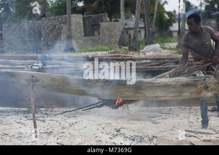 Fisherman utilizzando la masterizzazione di fronde di palma per rimuovere il muschio e verde crescite erbosa dal lato della sua nave da pesca in Jambiani Zanzibar Tanzania Foto Stock