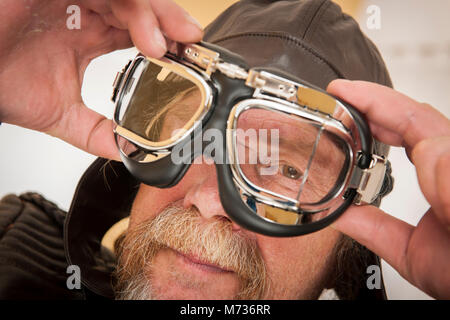 Close-up di un uomo anziano con la barba, Cappello in cuoio tiene con entrambe le mani una motocicletta occhiale davanti al suo volto e winks nella fotocamera Foto Stock