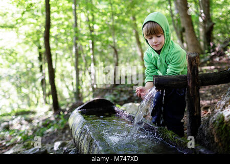Giovane ragazzo biondo acqua potabile dalle mani a tazza dal trogolo rustico in una foresta Foto Stock