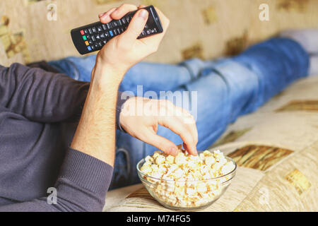 Un uomo con una ciotola di popcorn e un telecomando in mano guarda la TV sul divano Foto Stock