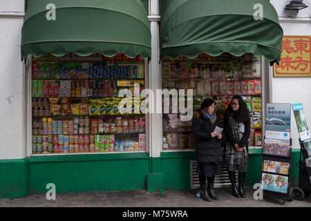 Due testimoni di Geova stand con copie della torre di avvistamento magazine accanto alla vetrina di un negozio di spuntini assortiti su scaffali in un angolo shop (convenience store) su Gerrard Street, Chinatown, il 5 marzo 2018, a Londra, in Inghilterra. Foto Stock