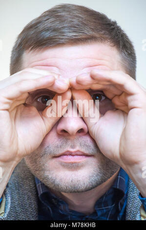 Ritratto di giovane divertente giocoso uomo tenendo le dita vicino agli occhi in una forma di immaginario binocolo su sfondo bianco. Foto Stock
