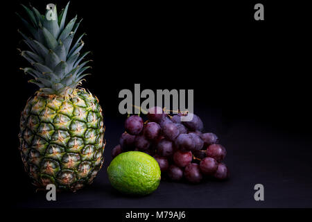 Cibo scuro - illuminazione in chiaroscuro su ananas, uve e calce con sfondo scuro e spazio copia a destra Foto Stock