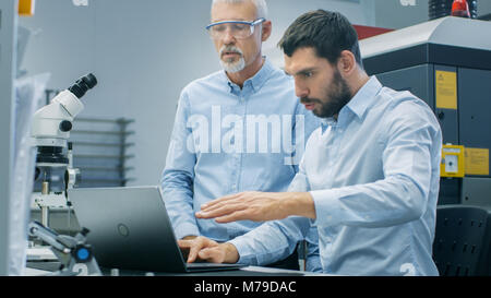 Due scienziati Industriale/ ingegneri/ tecnologi hanno discussione mentre si lavora sul computer portatile. In background moderno laboratorio/ Factory Foto Stock