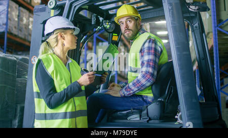 Female Supervisor Gives Work Assignment to Forklift Driver. They're in a Big Warehouse Full of Pallet Racks. Foto Stock