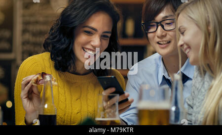 Nel Bar/Ristorante variegato gruppo di amici hanno un buon tempo, bello Uomo asiatico condivide foto e whatnot con il suo smartphone. Bella gente Foto Stock